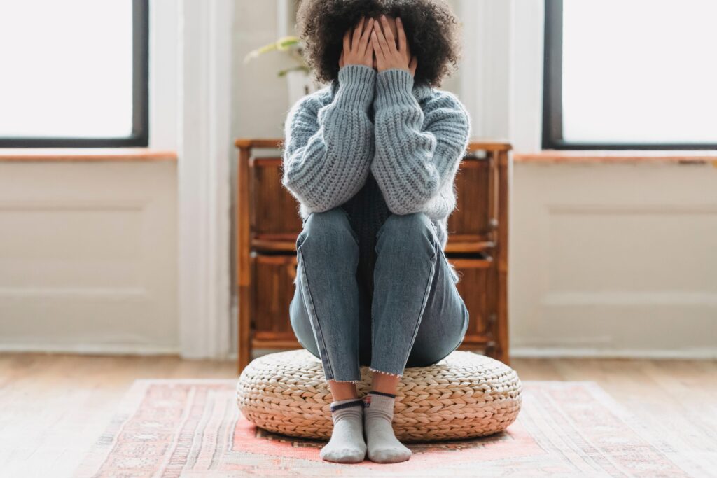 A woman with medium skin and curly dark brown hair sits on a round cushion on the floor with her elbows resting on her knees and her hands covering her face. The image suggests isolation and withdrawal as a stress response and accompanies a blog post about how distant leaders affect team psychological safety.