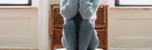 A woman with medium skin and curly dark brown hair sits on a round cushion on the floor with her elbows resting on her knees and her hands covering her face. The image suggests isolation and withdrawal as a stress response and accompanies a blog post about how distant leaders affect team psychological safety.