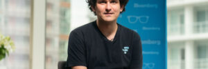 A photograph of Sam Bankman-Fried, founder of collapsed cryptocurrency exchange FTX. Bankman-Fried is seated wide-legged in front of a window through which skyscrapers can be seen. A blue banner, as used for an interview, is behind him. SBF gazes in the direction of the camera, and his hands are folded in his lap. He is a 28-year-old white man with unkempt, dark, curly hair. He is wearing a baggy black T-shirt and khaki cargo shorts. The photo accompanies a blog post about the personality of entrepreneurs and Bankman-Fried’s role in a particular kind of entrepreneurial culture.