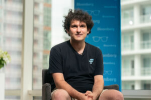 A photograph of Sam Bankman-Fried, founder of collapsed cryptocurrency exchange FTX. Bankman-Fried is seated wide-legged in front of a window through which skyscrapers can be seen. A blue banner, as used for an interview, is behind him. SBF gazes in the direction of the camera, and his hands are folded in his lap. He is a 28-year-old white man with unkempt, dark, curly hair. He is wearing a baggy black T-shirt and khaki cargo shorts. The photo accompanies a blog post about the personality of entrepreneurs and Bankman-Fried’s role in a particular kind of entrepreneurial culture.