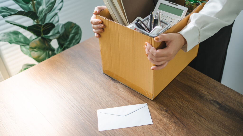 In this photograph, a person with tan-painted fingernails holds a yellow cardboard file box filled with office supplies. Only the person’s midbody is in the frame, wearing a long-sleeved white shirt and black slacks. The box is rested on the edge of a dark wood table. The box contents include writing utensils in a mesh cup, a calculator, notebooks, folders, and a potted plant. Next to the table is a large potted plant in front of a window covered with blinds. Atop the table is an envelope that presumably contains a resignation letter. The photo accompanies a blog post about how to resign and how not to resign, based on the approaches to resignation taken by Christine Lambrecht and Jacinda Ardern. The blog post explains how the two politicians’ ways of resigning differed in terms of personality and strategic self-awareness.