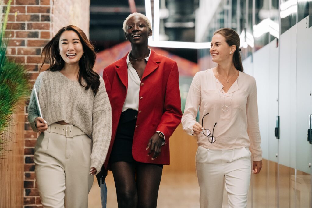 Three business colleagues of different races and ages walk together along a brick and glass hallway in a modern office. Their energetic postures and confident expressions suggest that they experience well-being in the workplace. The image accompanies a blog post about the importance of inclusion and belonging for well-being initiatives.