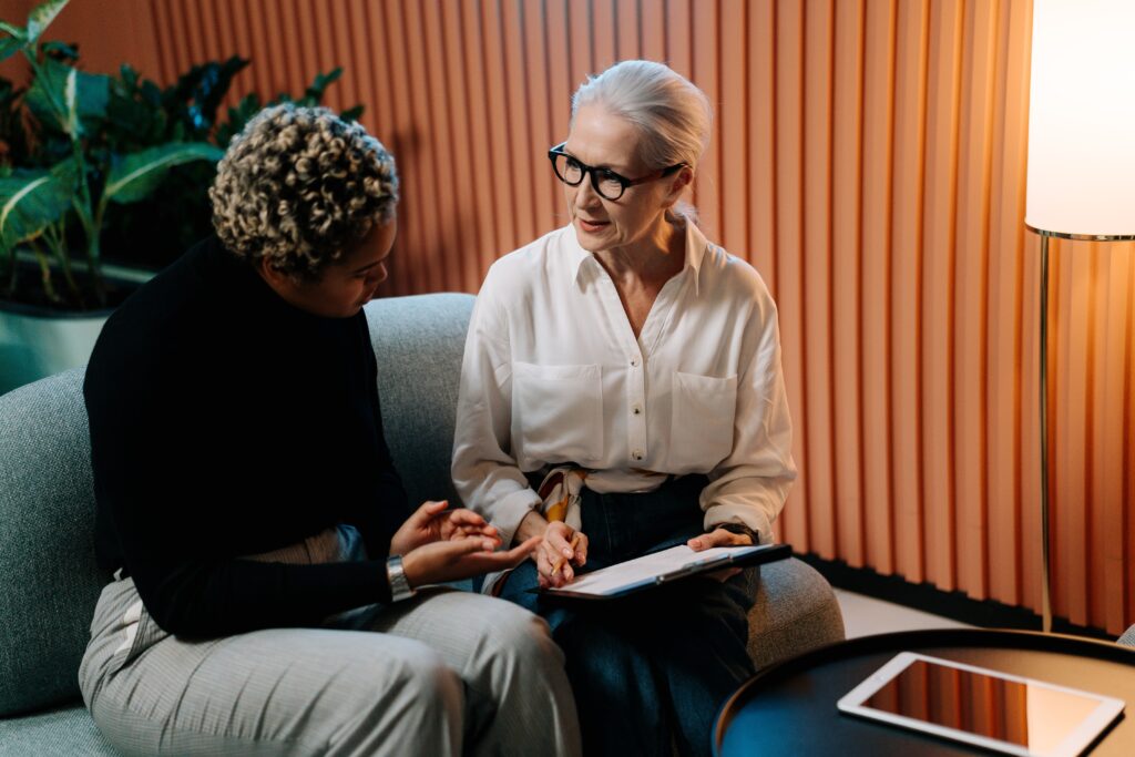 A white-haired person wearing glasses and a white blouse sits beside an early-career worker on a sofa discussing the contents of a clipboard. The two appear to be having a serious conversation, as an HR leader might have with an employee. The effect of HR burnout on the well-being of other employees is a main topic of the accompanying blog post.