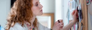 In this photo, a person with long, curly red hair and pale skin, who is wearing a loose white blouse and beaded jewelry, pins a photograph of a fashion model to a cork bulletin board. The board features various fashion sketches, photographs, and pages torn from magazines. It appears to be a vision board for goal setting. The room behind the person is out of focus, but a potted plant and a large mirror are visible against a white wall in the background. This photo accompanies a blog post about the relationship between personality and life goals.