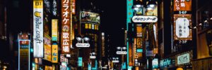 A photo of a street in Tokyo, Japan, taken at night. The street is crowded with people's silhouettes and illuminated with numerous neon lights. The photo accompanies a blog post about Hogan's recent trip to Tokyo to present at public events and visit with distributors, Optimal and Persol.