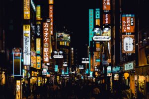 A photo of a street in Tokyo, Japan, taken at night. The street is crowded with people's silhouettes and illuminated with numerous neon lights. The photo accompanies a blog post about Hogan's recent trip to Tokyo to present at public events and visit with distributors, Optimal and Persol.
