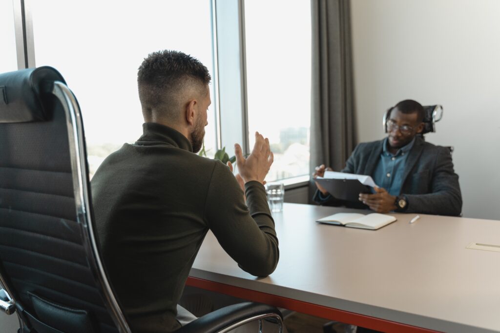 A business leader at a small conference table is conversing with an employee. The leader is consulting a clipboard of papers and has an open notebook and pen on the table. The employee gestures emphatically while speaking. It appears that they may be having a development conversation, and the employee may be responding with feedback resistance.