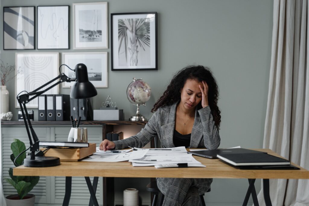 A medium-skinned person with long, dark, curly hair sits at a desk in an office resting their head on their hand with a weary expression. Their other hand rests on top of a slew of papers spread around the desktop. The desk also holds a closed laptop, a task lamp switched off, a cup of writing utensils, and some books. The person is wearing a black and white plaid suit with a black blouse underneath. Behind them is a gray wall, a file cabinet, some binders, a globe, and some wall art and other decor. A houseplant and some long gray curtains are also visible in the frame of the photo. The photo accompanies a blog post about incorporating well-being into leadership development discussions.