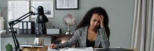 A medium-skinned person with long, dark, curly hair sits at a desk in an office resting their head on their hand with a weary expression. Their other hand rests on top of a slew of papers spread around the desktop. The desk also holds a closed laptop, a task lamp switched off, a cup of writing utensils, and some books. The person is wearing a black and white plaid suit with a black blouse underneath. Behind them is a gray wall, a file cabinet, some binders, a globe, and some wall art and other decor. A houseplant and some long gray curtains are also visible in the frame of the photo. The photo accompanies a blog post about incorporating well-being into leadership development discussions.