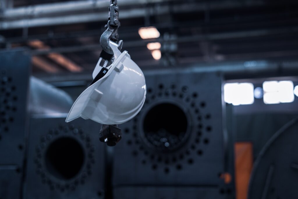 A white hard hat hangs from a steel hook in front of a dark industrial background. The photo accompanies a blog post about using personality assessments to create a safe workplace environment and save lives.