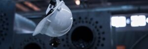 A white hard hat hangs from a steel hook in front of a dark industrial background. The photo accompanies a blog post about using personality assessments to create a safe workplace environment and save lives.