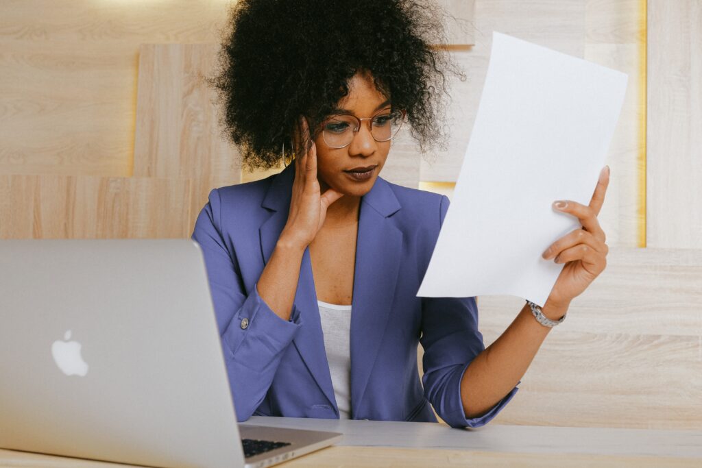 A person wearing glasses and a white blouse with a periwinkle blazer, who has curly dark hair and medium skin, rests one hand on her face and holds a document in the other. Her elbows are resting on the table as she reviews the document. A MacBook is open on the table in front of her. Behind her is a pale-colored contemporary wood-paneled wall. The image accompanies a blog about ethical considerations in workplace assessments. The implication is that the person in the photo is an IO psychologist or talent management professional who values ethics in workplace assessments.