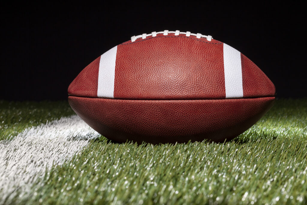 A low-angle photo of an American football on a grass field with a black background. A field yard line is visible just to the left of the ball. The photo accompanies a blog post about the similarities in leadership styles and personality between American football player turned coach Deion Sanders and Twitter CEO Elon Musk.