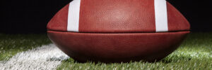 A low-angle photo of an American football on a grass field with a black background. A field yard line is visible just to the left of the ball. The photo accompanies a blog post about the similarities in leadership styles and personality between American football player turned coach Deion Sanders and Twitter CEO Elon Musk.