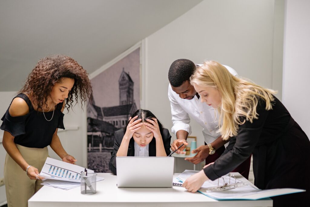 Four office workers appear to be frustrated and overwhelmed by collaborating with each other. One is sitting at a laptop holding their head in their hands, and the other three, standing, are leaning over them in postures of impatience or insistence. This image accompanies a blog post about change fatigue, team derailment, and versatile leadership.