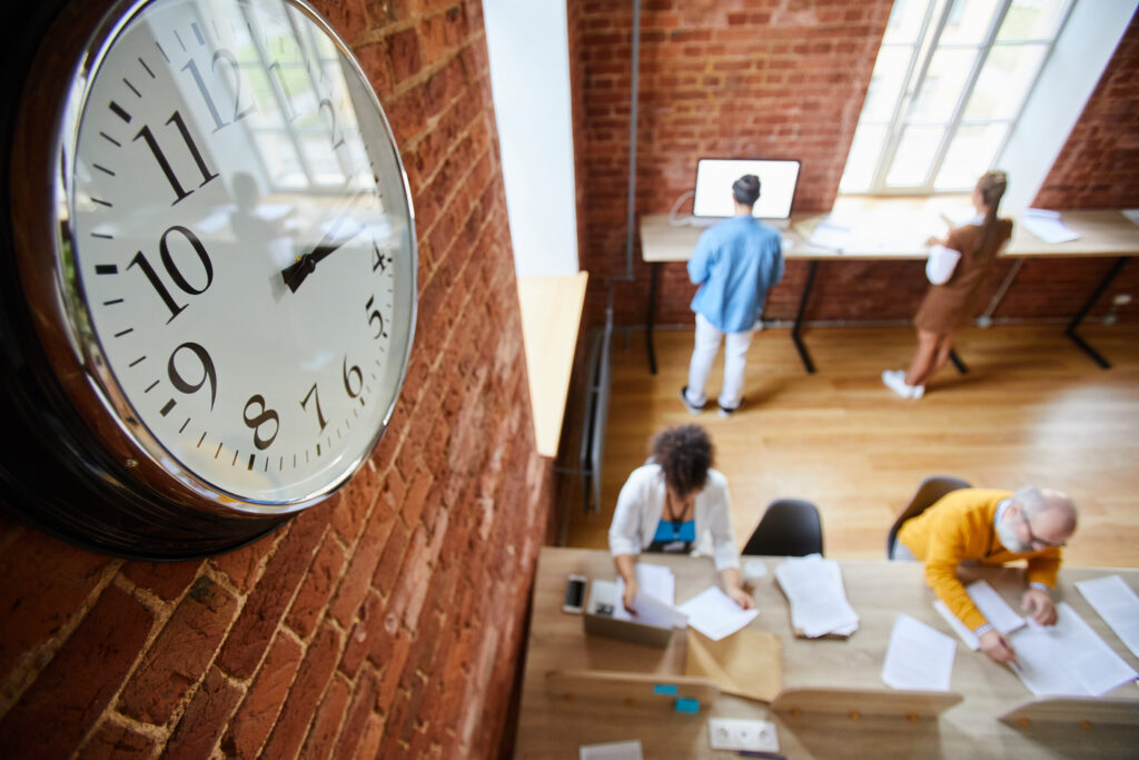 A bird's-eye view photo of an office space with brick walls, high ceilings, and tall windows through which bright light shines. A clock hangs on the wall in the left foreground of the photo. Below, a diverse group of employees are working at long tables on opposite sides of the room. The photo accompanies a blog post about soft skills or socioemotional skills having a "moment" in the literature, although these skills have always been important for leaders and talent in general.