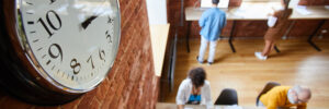 A bird's-eye view photo of an office space with brick walls, high ceilings, and tall windows through which bright light shines. A clock hangs on the wall in the left foreground of the photo. Below, a diverse group of employees are working at long tables on opposite sides of the room. The photo accompanies a blog post about soft skills or socioemotional skills having a "moment" in the literature, although these skills have always been important for leaders and talent in general.