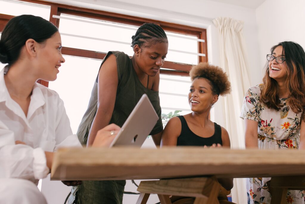 A diverse group of women are gathered around a conference table. One is working on a laptop, while the others appear to be reviewing papers not visible in the frame. A window with the curtains and blinds open is behind them. The photo accompanies a blog post about empowering the next generation of women leaders.