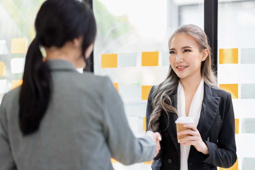 Two women in business attire shake hands in a conference room. One has her back to the camera; the other is facing the camera, smiling, and holding a paper cup of coffee. The photo accompanies a blog post about coaching leaders through change, which provides advice on leadership development and change management.