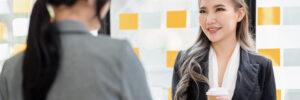 Two women in business attire shake hands in a conference room. One has her back to the camera; the other is facing the camera, smiling, and holding a paper cup of coffee. The photo accompanies a blog post about coaching leaders through change, which provides advice on leadership development and change management.