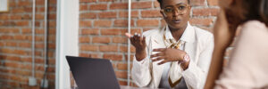 A dark-skinned person with short natural hair gestures while speaking to a medium-skinned person with long, straight brown hair who is out of focus and facing away from the camera at the right of the frame. The dark-skinned person is wearing an ivory blazer over a white button-down shirt with a silk scarf, a rose-gold wristwatch, orange- or pink-tinted clear acrylic eyeglasses, and light blue fingernail polish. In front of them, a laptop and a glass of water sit on a white or gray conference table surface. The other person is wearing a long-sleeved peach blouse with the sleeves rolled up. They are resting their arms on the table. In one hand, they are holding a piece of paper. Their chin rests in their other hand as they look at the person speaking. Behind the person speaking is a brick wall. The photo accompanies a blog post about redesigning annual performance review conversations, so the implication is that the two people featured are engaged in a performance appraisal.