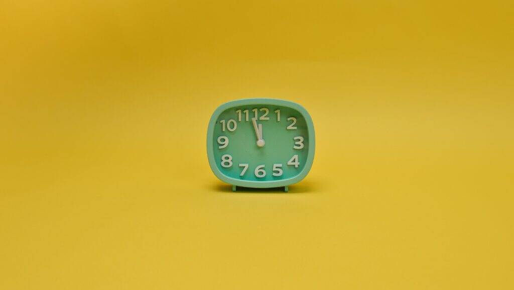 An analog clock against a canary yellow background. The clock is mint green with white numerals. Its hour arm is pointed at 12, and its minute arm is pointed betwee 11 and 12. It has no arm to measure seconds. The image accompanies a blog post about the psychology of patience and personality.  