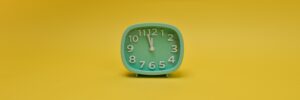 An analog clock against a canary yellow background. The clock is mint green with white numerals. Its hour arm is pointed at 12, and its minute arm is pointed betwee 11 and 12. It has no arm to measure seconds. The image accompanies a blog post about the psychology of patience and personality.