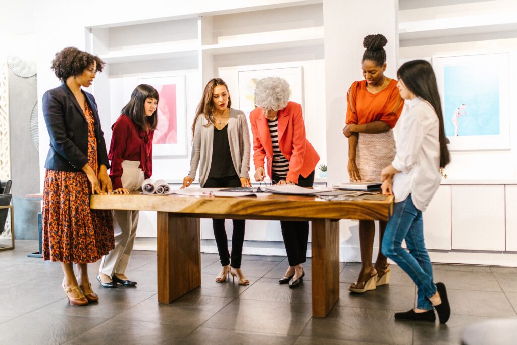 A diverse group of people stand around a wooden conference table, upon which they are reviewing several large pieces of paper. They appear to be working on a creative project together. The image accompanies a blog post about managing creative teams and competencies for leaders of creatives.
