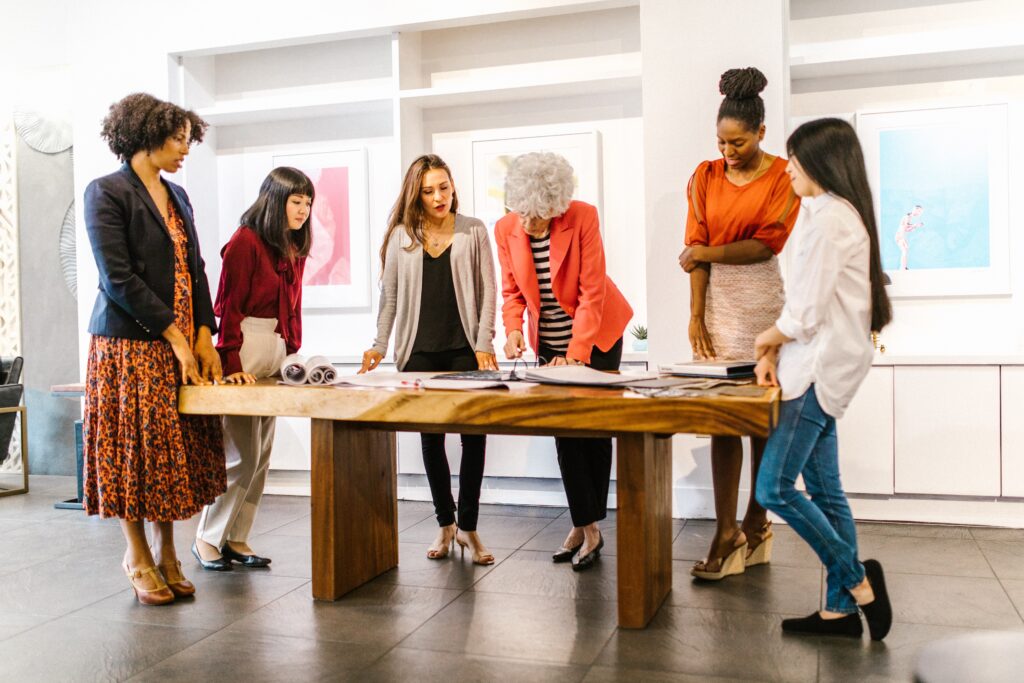 A diverse group of people stand around a wooden conference table, upon which they are reviewing several large pieces of paper. They appear to be working on a creative project together. The image accompanies a blog post about managing creative teams and competencies for leaders of creatives. 