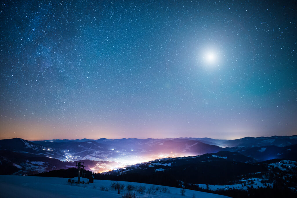 A landscape of a wintry mountain range against a starry night sky with a bright full moon. Amid the mountains is a glowing light, conceptualizing the mysticism of UFOs and abnormal phenomena. 