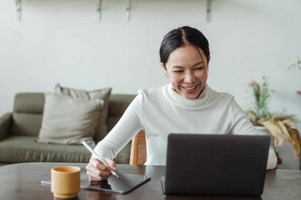 An olive-skinned person with black hair is wearing a long-sleeved ivory turtleneck. Their hair is tied up in a low updo. The person is seated at a wooden table in front of a laptop computer. The person is smiling broadly at the laptop while holding a digital pen over a digital tablet at their right side. They have a small tan mug near their tablet. In the background, a dull green couch with gray throw pillows sits against a white wall. Also against the wall is a dying fern houseplant. The photo accompanies a blog post about maximizing a talent development budget using personality assessments, so the person could be either planning talent development strategy or partaking in a talent development or leadership development initiative, such as coaching.