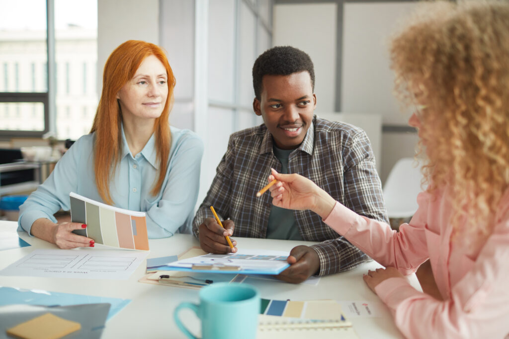 Three people discuss a creative project while seated around an off-white office table. At the right, a medium-skinned person with long coiled light brown hair and a pink blouse gestures to the other two with a pencil in their hand. The other two people look at the speaker and are smiling slightly. The dark-skinned person in the center has short hair and is wearing a brown plaid shirt unbuttoned over a green tee-shirt. They are holding a pencil and clipboard. Pictured at left, a light-skinned person with long straight copper hair and a light blue blouse is holding color swatches with their arm rested atop the table. Various color swatches, paper, other work materials, and a turquoise coffee cup also sit on the table.