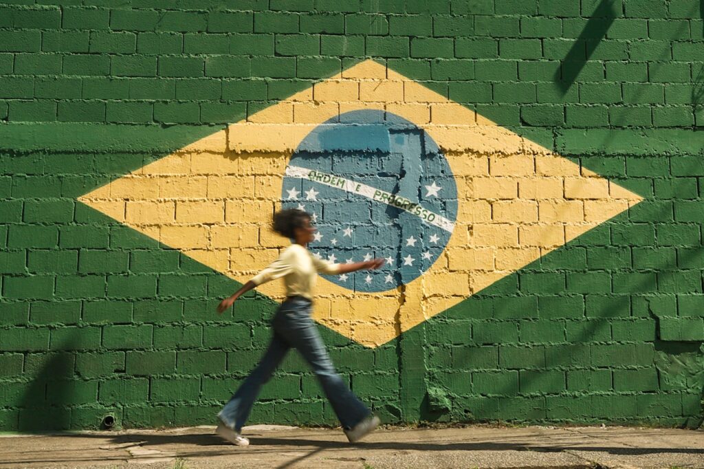 A person walks past a cinderblock wall painted to resemble the flag of Brazil. The mural-sized green, gold, blue, and white flag reads Ordem e Progresso in all capital letters. The person walking by is slightly out of focus and their arms and legs are at a wide stance, as if they are moving at a fast pace. They have medium skin and long dark hair that is pulled back, and they are wearing jeans, a white shirt, and white shoes. The atmosphere of the photo is sunny. The photo accompanies a blog post about the personality characteristics of Brazilian managers.