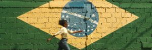A person walks past a cinderblock wall painted to resemble the flag of Brazil. The mural-sized green, gold, blue, and white flag reads Ordem e Progresso in all capital letters. The person walking by is slightly out of focus and their arms and legs are at a wide stance, as if they are moving at a fast pace. They have medium skin and long dark hair that is pulled back, and they are wearing jeans, a white shirt, and white shoes. The atmosphere of the photo is sunny. The photo accompanies a blog post about the personality characteristics of Brazilian managers.