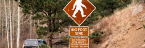 A brown and white roadside sign cautions passersby about potential bigfoot crossing. The sign notes that sightings of a “creature resembling ‘big foot’” have happened in the area and features a stereotypical Sasquatch silhouette (i.e., a hairy hominid figure). The sign is backed by a forested landscape with both bare trees and pine trees. The mountainside at the forefront of the image is sandy and stark with dry brush. On the road to the left of the sign, a bumper-stickered green SUV drives off. The photo accompanies a blog post about cryptozoology, the personality characteristics of cryptozoologists, and evidence of bigfoot.