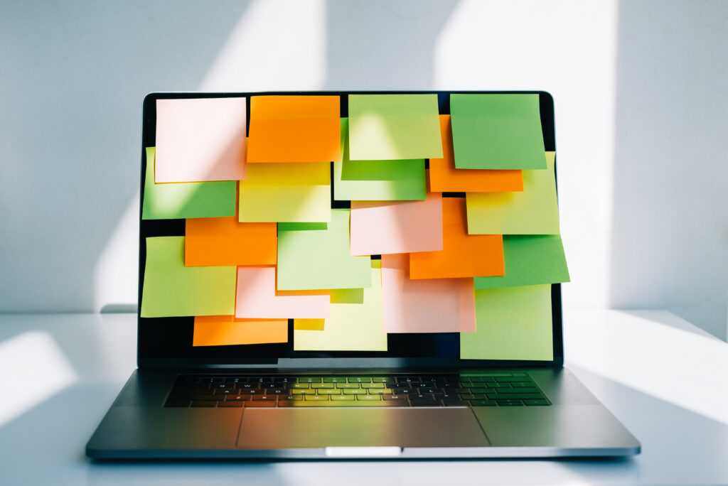 A MacBook laptop sits on a white desktop against a white wall. Its screen is covered from top to bottom and side to side with blank sticky notes in pink, orange, green, and yellow. The image accompanies a blog post about causes of employee burnout, individual differences in burnout, and how to manage employee burnout.