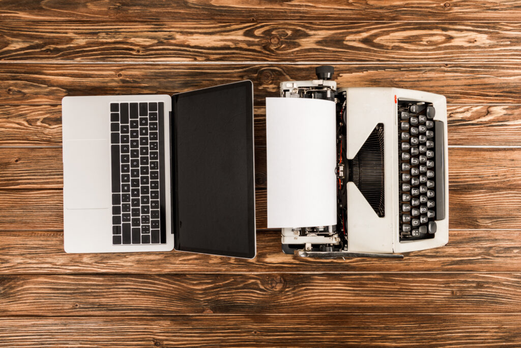 A top-down view of a typewriter and a laptop situated back-to-back on a wooden table. The two represent the transition from analog to digital to accompany an article about generational differences in personality.