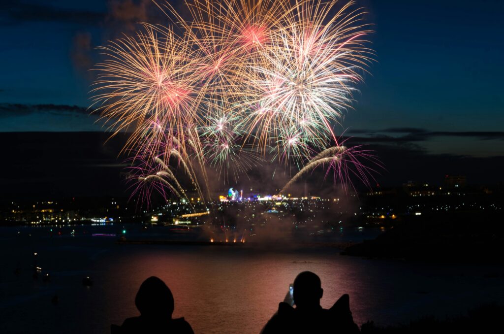 Fireworks explode over a cityscape and a body of water at nighttime. In the foreground of the photo are two human silhouettes. One of the people is using a smartphone to take a photo. Signifying the new year, the photo accompanies a blog post about the derailers that defined the year 2023.