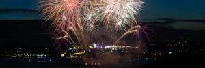 Fireworks explode over a cityscape and a body of water at nighttime. In the foreground of the photo are two human silhouettes. One of the people is using a smartphone to take a photo. Signifying the new year, the photo accompanies a blog post about the derailers that defined the year 2023.