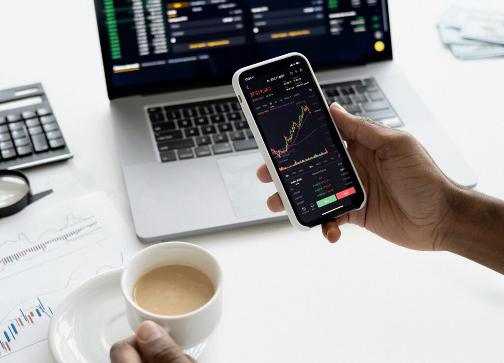 An investor reviews their investment data using a smartphone in one hand while holding a cup of coffee in the other. In front of them is a laptop displaying additional data.