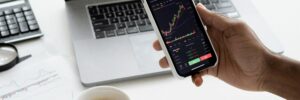 An investor reviews their investment data using a smartphone in one hand while holding a cup of coffee in the other. In front of them is a laptop displaying additional data.