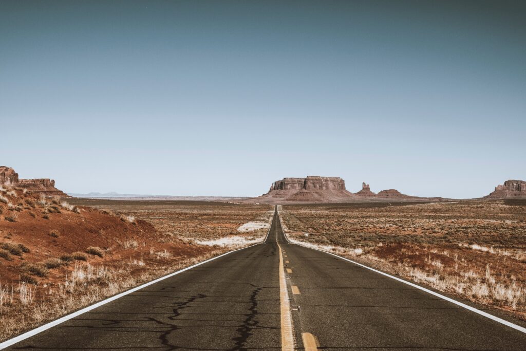 A two-lane highway, centered in the photo, stretches into an expansive desert landscape toward a couple of mesas and a butte against a cloudless sky. Additional formations are visible in the periphery. The photo accompanies a blog about how the entrepreneurial economy is a new frontier but has a dark side in terms of the personality characteristics it may expose.