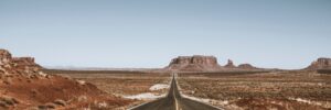 A two-lane highway, centered in the photo, stretches into an expansive desert landscape toward a couple of mesas and a butte against a cloudless sky. Additional formations are visible in the periphery. The photo accompanies a blog about how the entrepreneurial economy is a new frontier but has a dark side in terms of the personality characteristics it may expose.