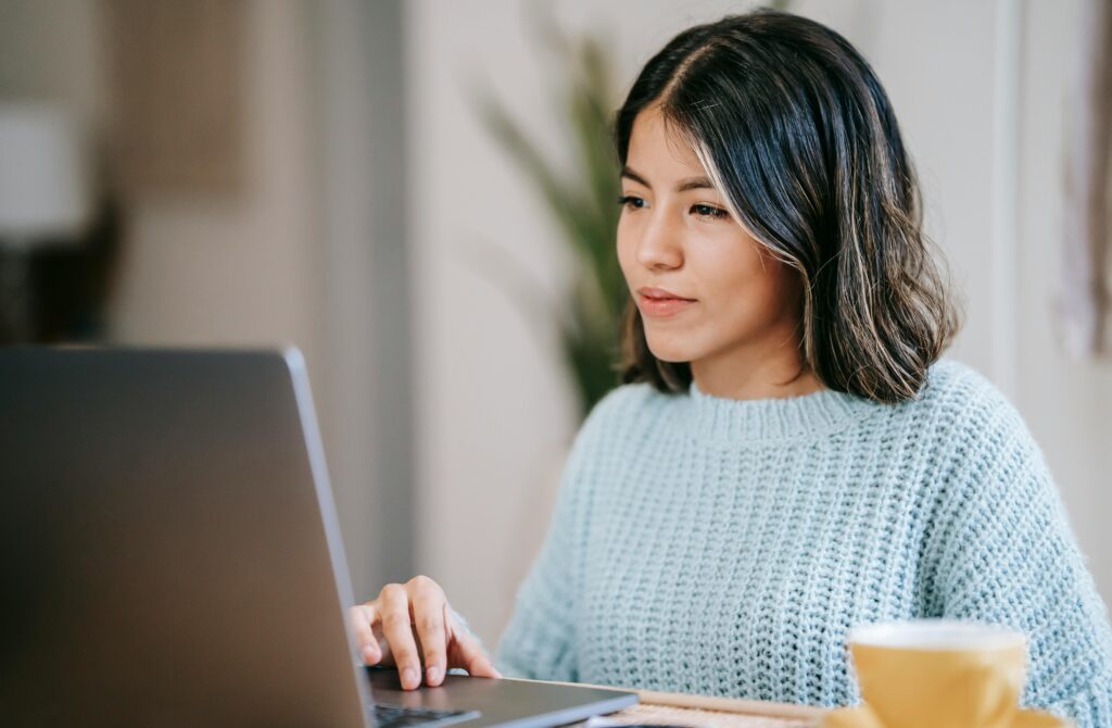 A medium-skinned person with shoulder-length dark hair uses a laptop. The person is wearing a gray sweater and has a tan coffee cup on a saucer at their left. Out of focus in the background is a plant. The photo accompanies a blog post about how to take a personality test.