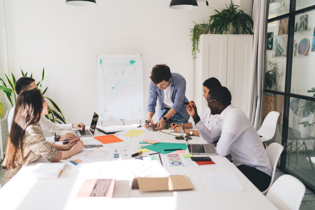 A multiethnic and multigendered team of professionals are sitting in white chairs around a white table, topped with papers, laptops, writing utensils, and smartphones while collaborating on a creative project. The photo accompanies a blog post about the importance of values, which notes that cooperation is a value important to organizational success.