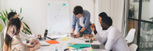 A multiethnic and multigendered team of professionals are sitting in white chairs around a white table, topped with papers, laptops, writing utensils, and smartphones while collaborating on a creative project. The photo accompanies a blog post about the importance of values, which notes that cooperation is a value important to organizational success.