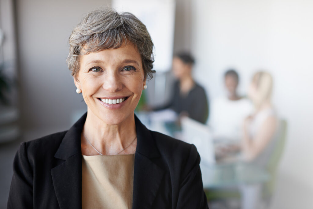 A person with short gray hair wearing a black blazer over a gold blouse smiles at the camera. The person is wearing pearl earrings and a silver necklace. In the background, other professionals are visible but out of focus. The photo accompanies a blog post about the metacompetencies of leadership.