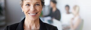 A person with short gray hair wearing a black blazer over a gold blouse smiles at the camera. The person is wearing pearl earrings and a silver necklace. In the background, other professionals are visible but out of focus. The photo accompanies a blog post about the metacompetencies of leadership.