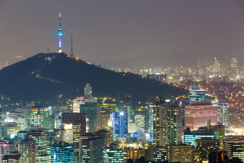 A panoramic view of the Seoul skyline at night features N Seoul Tower glowing blue atop Namsan Mountain above illuminated skyscrapers. The image of the vibrant South Korean urban landscape accompanies an article about leadership insights of South Korean leaders.