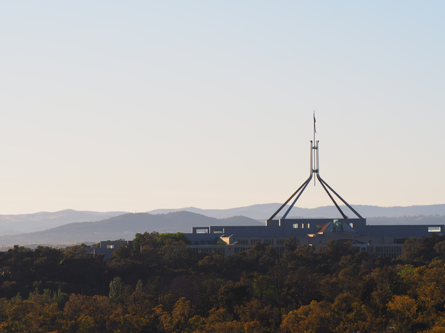 Parliament House sits prominently on Capital Hill in Canberra, Australia, its distinctive flagpole rising above the modern building’s angular roofline against a backdrop of rolling hills. The image accompanies a profile of the personality characteristics of Australian leaders and business executives.